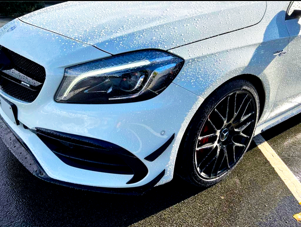 A white car, with water droplets on its surface, is parked in a lot. The vehicle features a sleek front design, prominent headlights, and shiny black alloy wheels.