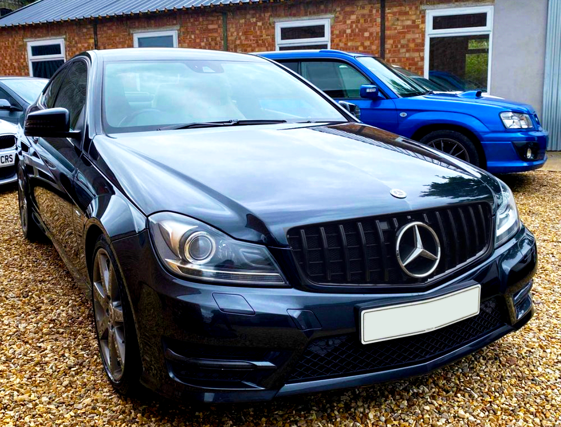 A black Mercedes-Benz sedan is parked on a gravel lot, surrounded by other vehicles in front of a brick building.