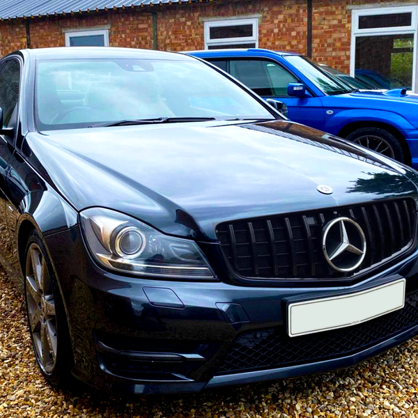 A black Mercedes-Benz sedan is parked on a gravel lot, surrounded by other vehicles in front of a brick building.