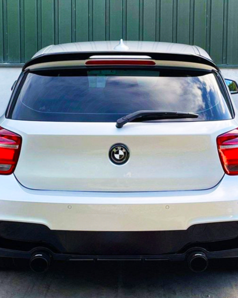 A white hatchback car, with a BMW logo, is parked in front of a green metal wall. It features a rear wiper and dual-exhaust pipes. Reflective glass shows clouds.
