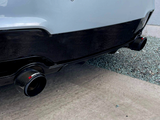 Exhaust pipes with carbon fiber tips are affixed to the rear of a car, positioned above gravel and pavement. The text 