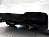 A white car's rear bumper with a black diffuser features dual exhaust pipes, located indoors, with another red vehicle partially visible in the background.