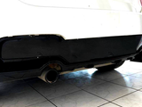 A black diffuser with dual exhaust pipes is attached to a white car, parked indoors on a tiled floor next to a blue vehicle.