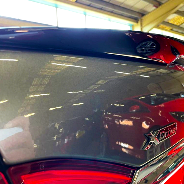 Close-up of a car's sleek, reflective surface displaying the "XDrive" badge, surrounded by reflections of red cars in a spacious, industrial garage with a high ceiling.