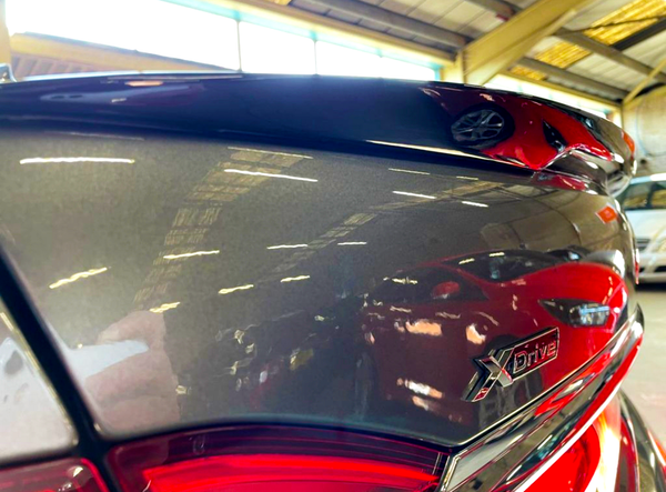Close-up of a car's sleek, reflective surface displaying the "XDrive" badge, surrounded by reflections of red cars in a spacious, industrial garage with a high ceiling.