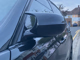 A black car's side mirror captures reflections of houses, parked on a suburban street lined with residential buildings and trees, during dusk.
