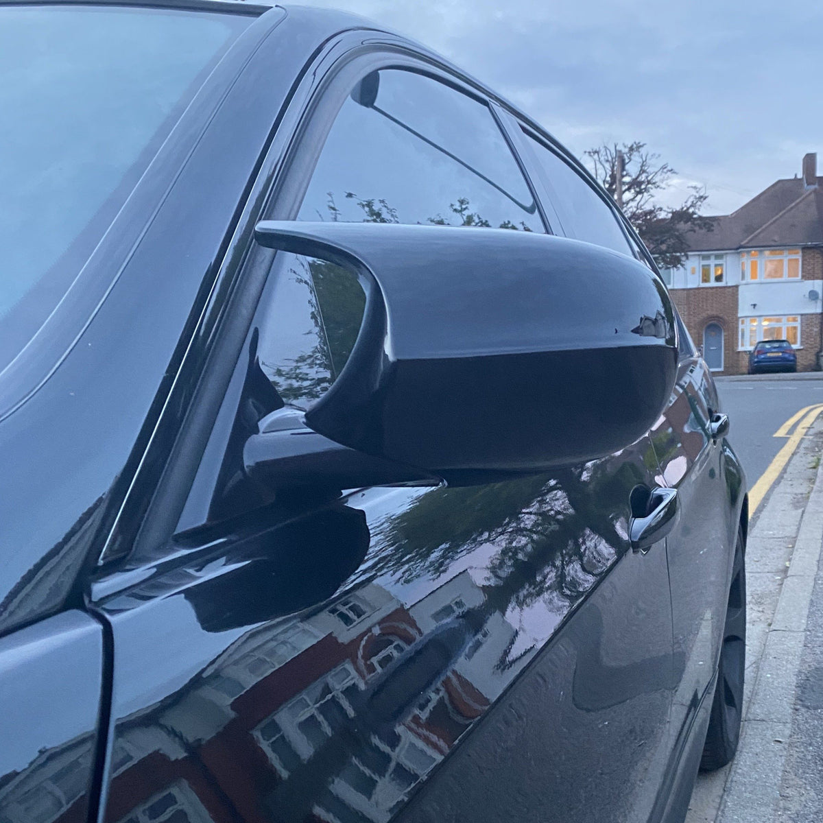 A black car's side mirror captures reflections of houses, parked on a suburban street lined with residential buildings and trees, during dusk.