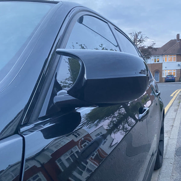 A black car's side mirror captures reflections of houses, parked on a suburban street lined with residential buildings and trees, during dusk.