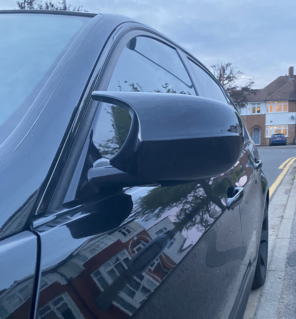 A black car's side mirror captures reflections of houses, parked on a suburban street lined with residential buildings and trees, during dusk.