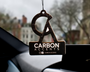 An air freshener hangs from a car's rearview mirror. It bears the text "CARBON ACCENTS" and social media tag "@carbon.accents." The windshield view shows blurred city buildings and trees.