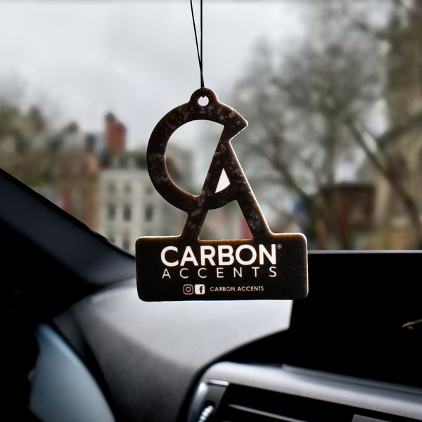 An air freshener hangs from a car's rearview mirror. It bears the text "CARBON ACCENTS" and social media tag "@carbon.accents." The windshield view shows blurred city buildings and trees.