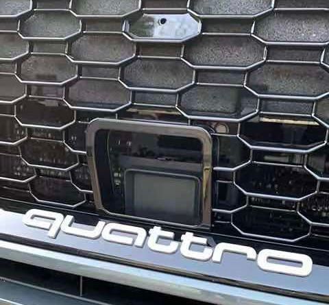 A car grille features a prominent honeycomb pattern. Centered is a rectangular sensor or camera. Below, bold white lettering spells "quattro," indicating a four-wheel drive system. The setting suggests an outdoor environment.
