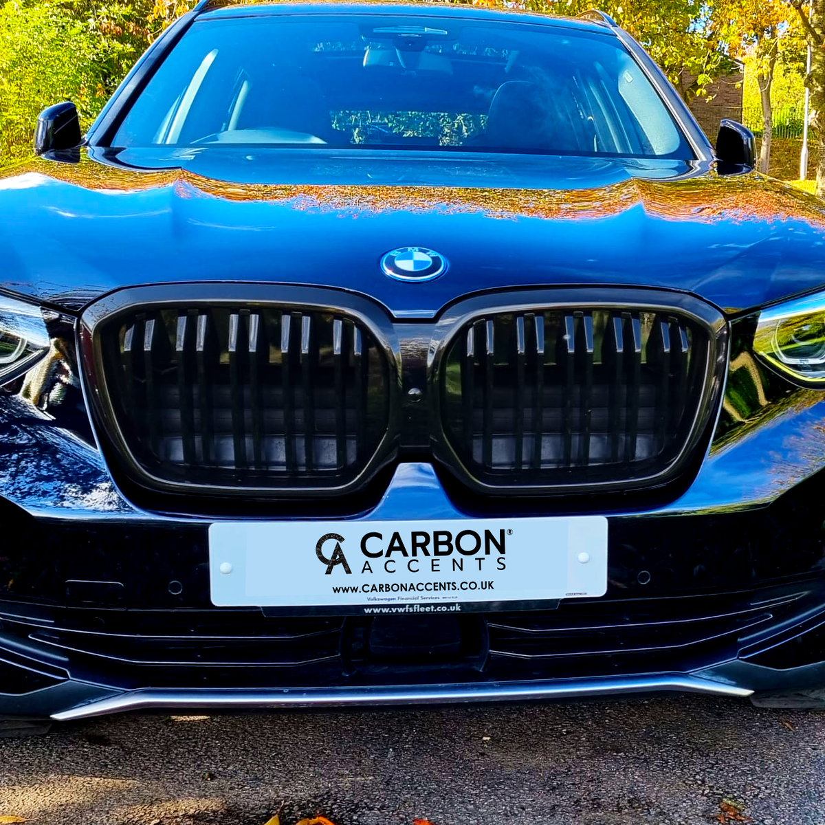 A black BMW SUV is parked on a sunlit road, surrounded by trees. The front license plate reads: "CARBON ACCENTS" with websites "CARBONACCENTS.CO.UK" and "www.rvsfleet.co.uk."