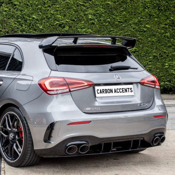 A sleek silver hatchback car is parked on a concrete driveway, featuring a prominent rear spoiler and red brake calipers. "CARBON ACCENTS" is inscribed on the license plate, adjacent to a lush green hedge.