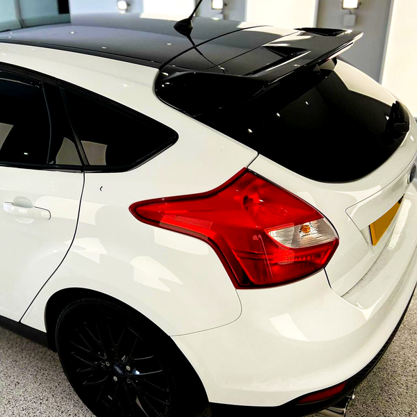 A white car with a black roof and spoiler, parked indoors on a speckled floor. It features a tinted rear window and red taillights. The setting is well-lit.