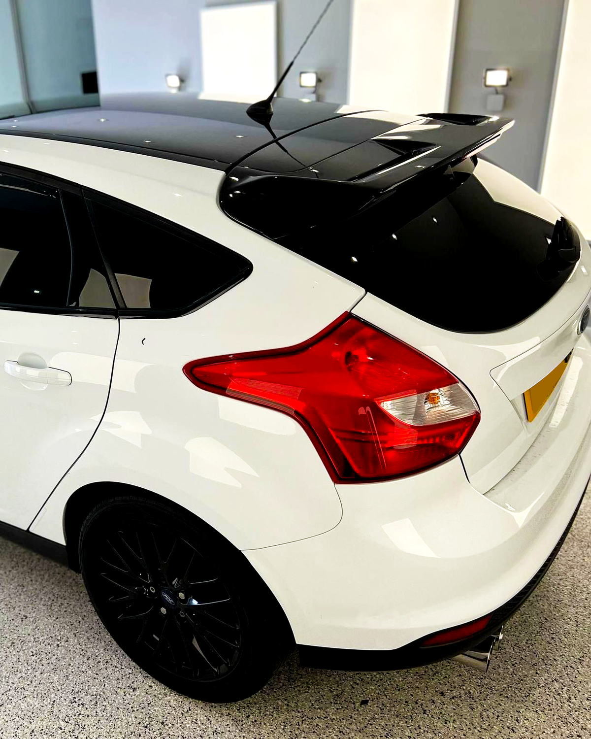 A white car with a black roof and spoiler, parked indoors on a speckled floor. It features a tinted rear window and red taillights. The setting is well-lit.