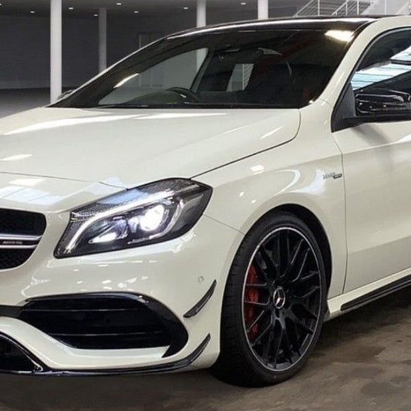 A white car with illuminated headlights is parked in an indoor showroom. It has sporty black rims and visible branding on the front. The plate reads "CARBON ACCENTS."