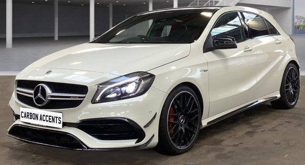 A white car with illuminated headlights is parked in an indoor showroom. It has sporty black rims and visible branding on the front. The plate reads "CARBON ACCENTS."