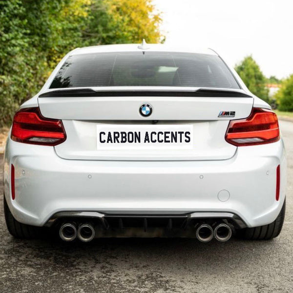 A white car, viewed from behind, displaying "CARBON ACCENTS" on its license plate, is parked on a road surrounded by greenery and trees.