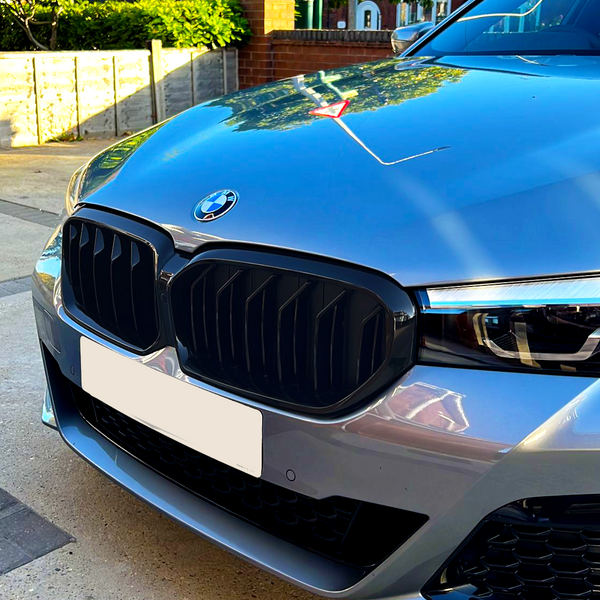 A silver BMW car is parked on a driveway, with a prominently visible grille and headlight. Trees and brick houses form the background under a clear blue sky.