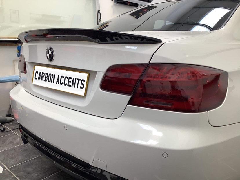 A white BMW is parked indoors, showcasing a carbon fiber spoiler. The license plate reads "CARBON ACCENTS." The environment features a tiled floor and white walls.
