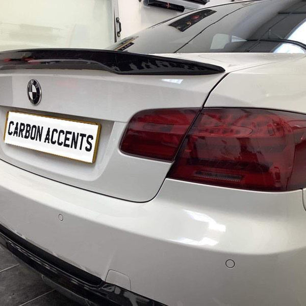 A white BMW is parked indoors, showcasing a carbon fiber spoiler. The license plate reads "CARBON ACCENTS." The environment features a tiled floor and white walls.