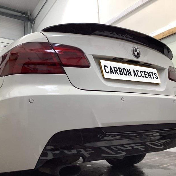 A white car's rear view in a garage displays a custom license plate with "CARBON ACCENTS" text. The context includes visible wall shelving with items like a spray bottle.