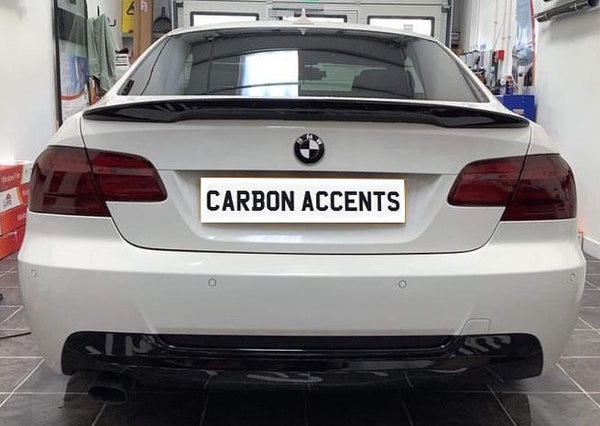 A white BMW is parked in a garage. It features a "CARBON ACCENTS" license plate. The setting includes tools and equipment on shelves.