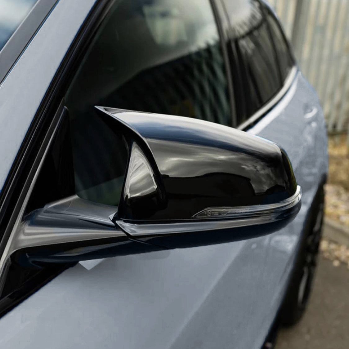 A shiny black side mirror is attached to a sleek gray car, reflecting clouds, with a blurred urban background featuring a metal fence and pavement.