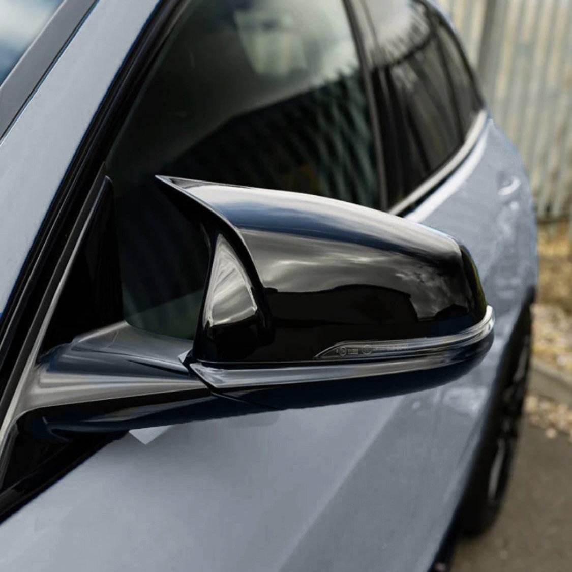 A shiny, black car side mirror reflects nearby surroundings, attached to a sleek blue vehicle parked on a paved area with metallic fencing in the background.