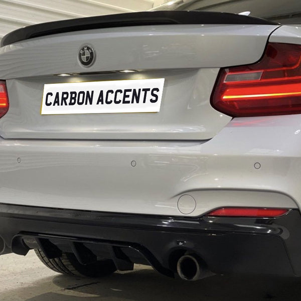 A white car with illuminated taillights, featuring dual exhausts and a sleek carbon diffuser, is parked indoors. License plate text reads: "CARBON ACCENTS."