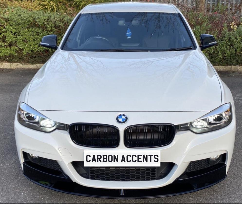 A white BMW car is parked on a paved surface, with "CARBON ACCENTS" text on the grille. Surrounding greenery and a fence are in the background.