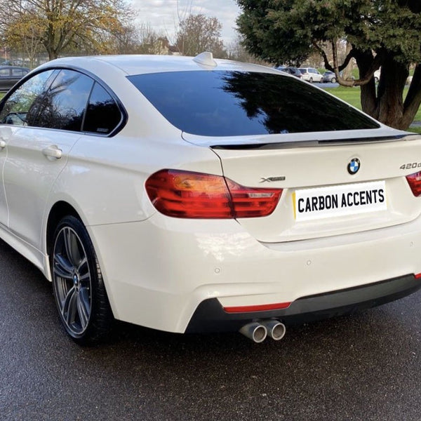 A white BMW 420d is parked on a paved road by a grassy area with trees. The license plate reads "CARBON ACCENTS." Several people and cars are visible in the background.