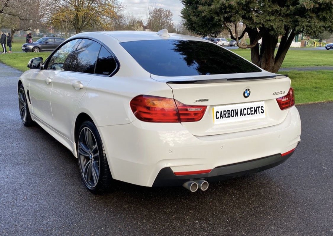 A white BMW 420d is parked on a paved road by a grassy area with trees. The license plate reads "CARBON ACCENTS." Several people and cars are visible in the background.