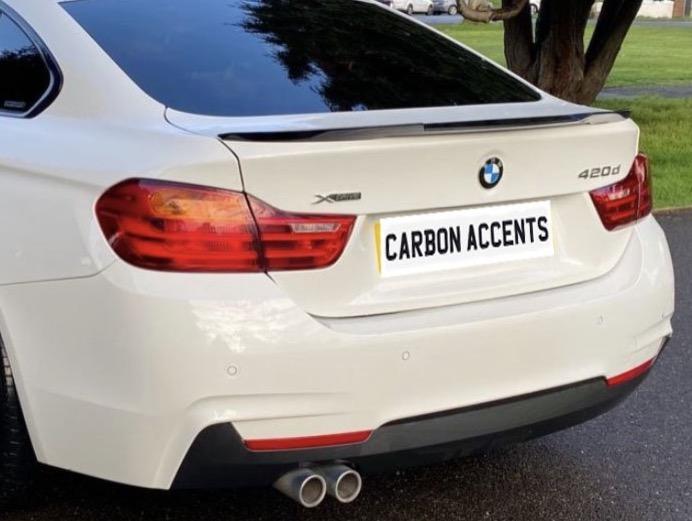 A white BMW 420d with tinted windows is parked on a wet pavement. The car features dual exhausts, red tail lights, and a visible license plate reading "CARBON ACCENTS."