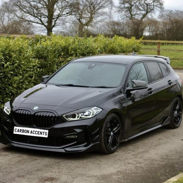A black car is parked on a road surrounded by greenery and trees. The front license plate reads "CARBON ACCENTS."