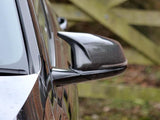 Car side mirror with carbon fiber casing reflecting the car's sleek body, set against a blurred wooden fence background.