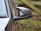 A carbon-fiber car side mirror reflects a blurred background of a wooden fence and greenery, attached to the sleek side panel of a dark-colored vehicle.