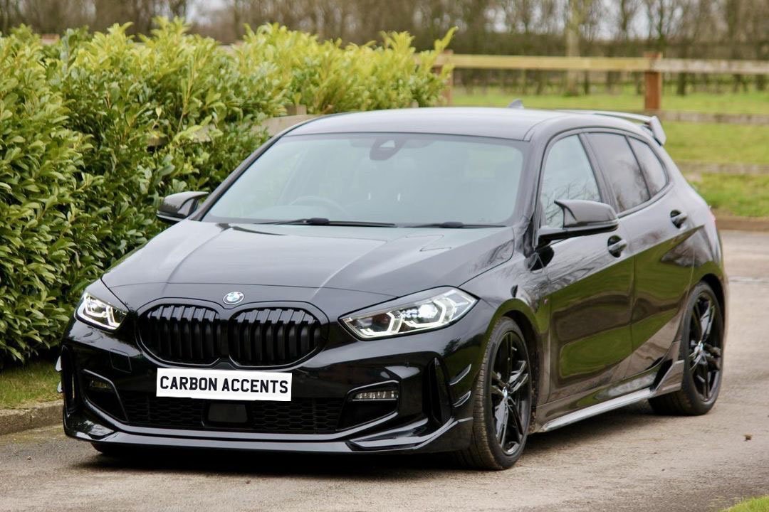 A black car is parked on a driveway, surrounded by greenery. The license plate reads "CARBON ACCENTS."
