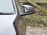 A car's glossy black side mirror is angled outward, reflecting the adjacent environment. The background reveals a gravel driveway and a wooden fence in an outdoor setting.