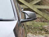 A black car's side mirror reflects a wooden gate in a gravel and grass driveway. The mirror has a carbon fiber texture and contrasts the blurred rustic background.