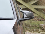 A black car with a carbon fiber side mirror is parked, facing a wooden fence in a gravel area.