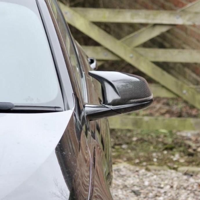 A black car with a carbon fiber side mirror is parked, facing a wooden fence in a gravel area.