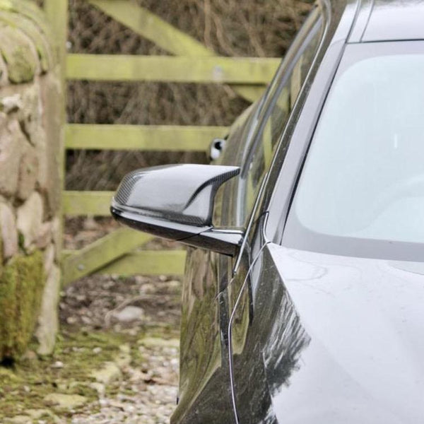 A sleek car's side mirror is shown outside near a moss-covered stone wall and a wooden gate in a rural setting.
