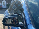 A blue car's side mirror reflects brick houses, parked on a residential street with sunlight highlighting the vehicle's metallic paint and nearby suburban homes.