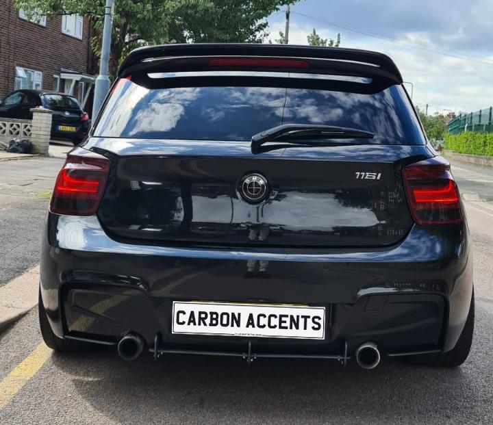 A black car with a clean rear design is parked on a street. The license plate reads "CARBON ACCENTS." Surrounding residential homes and trees create a suburban setting.