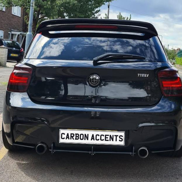A black car with a clean rear design is parked on a street. The license plate reads "CARBON ACCENTS." Surrounding residential homes and trees create a suburban setting.