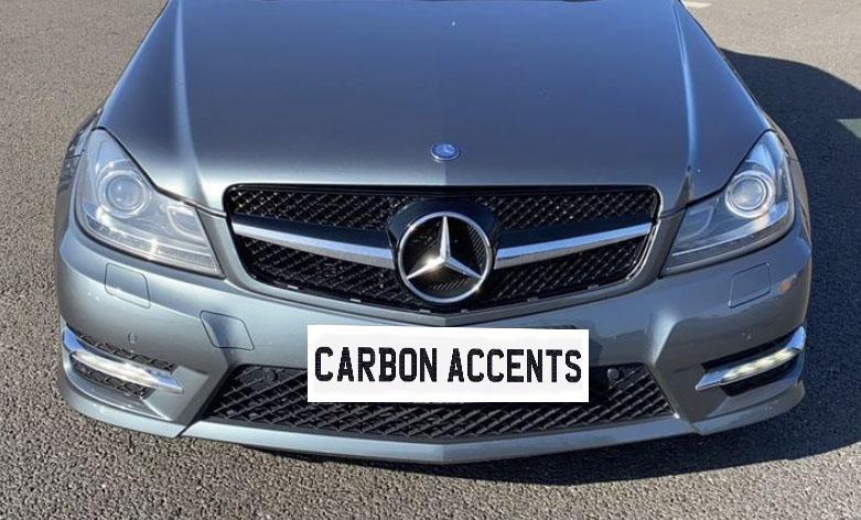 Silver car front, featuring a Mercedes-Benz emblem on the grille, parked on asphalt. Text on bumper reads "CARBON ACCENTS."