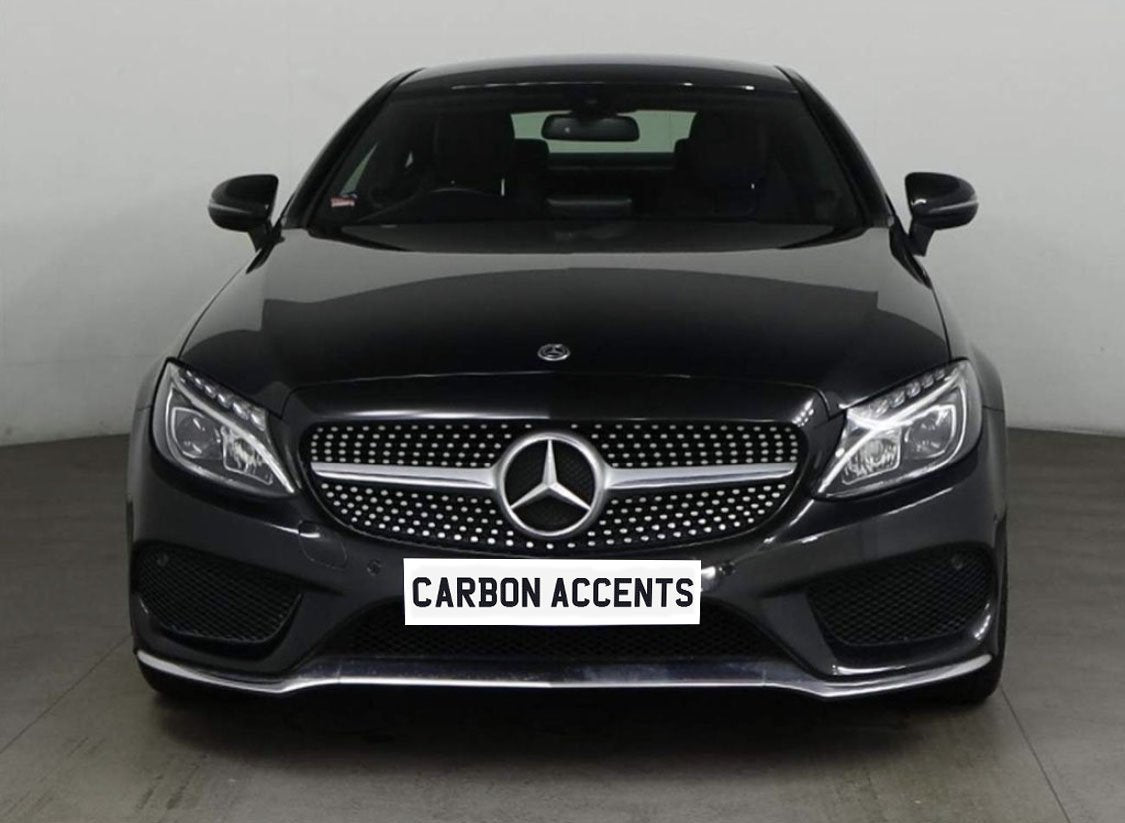 A black Mercedes-Benz car is parked indoors, facing front. Its grille features the Mercedes emblem. Below, a plate displays the text "CARBON ACCENTS." The background is plain white.