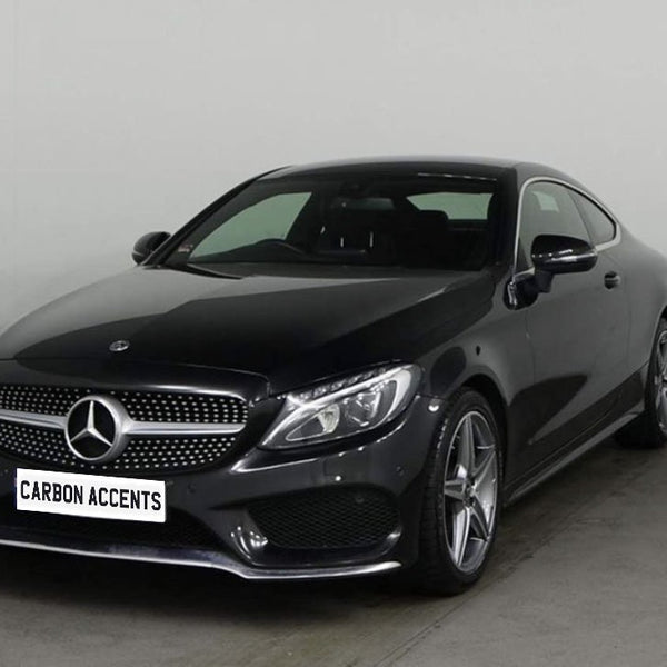 A sleek black Mercedes-Benz coupe is parked in a plain, well-lit showroom. The front license plate reads "CARBON ACCENTS," and the car features shiny alloy wheels.